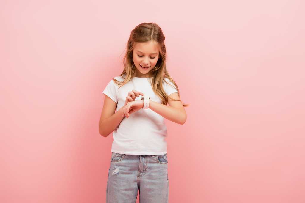 dziecko w białej koszulce i dżinsach dotykając smartwatch izolowany na różowy - Zdjęcie, obraz