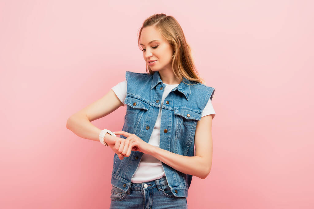 молода жінка в джинсовому жилеті, торкаючись смарт-годинника, ізольована на рожевому
 - Фото, зображення