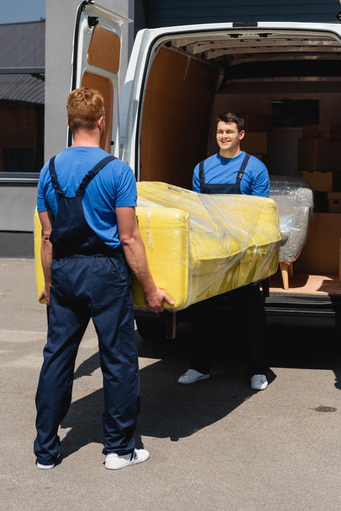Szelektív fókusz rakodó szállító kanapé nyújtózkodó csomagolásban kollégájával teherautó közelében a városi utcán  - Fotó, kép