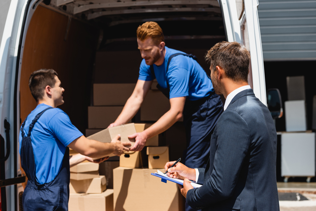 Selective focus of businessman holding pen and clipboard while movers unloading packages from truck outdoors  - Photo, Image