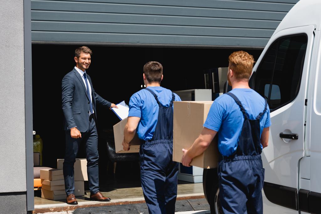 Concentration sélective de l'homme d'affaires tenant presse-papiers tandis que les déménageurs transportant des colis près du camion et de l'entrepôt à l'extérieur - Photo, image