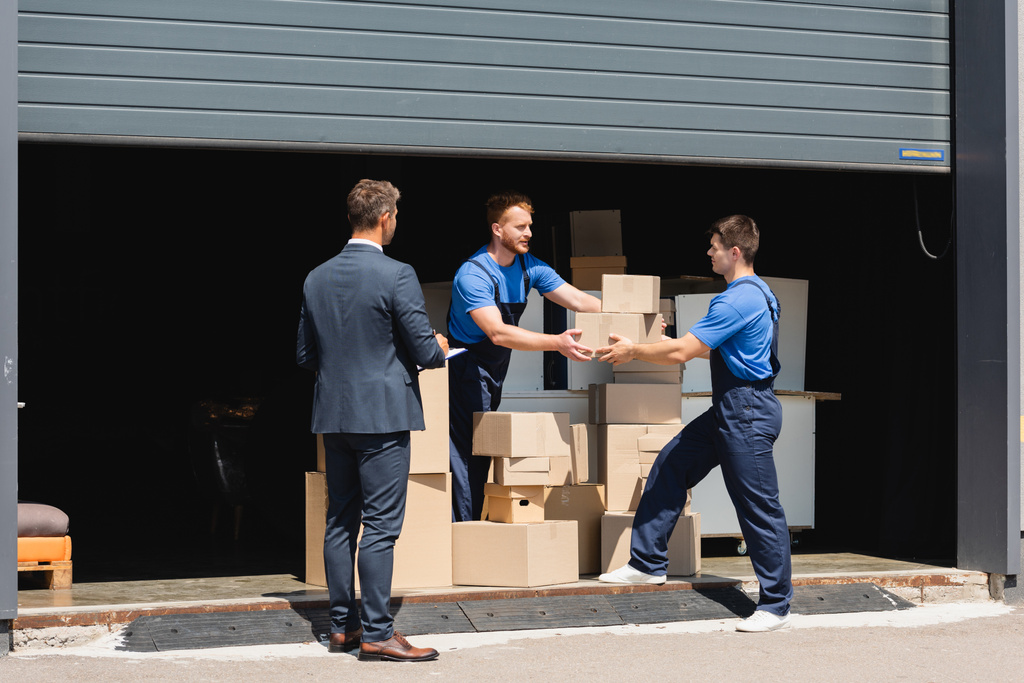Επιχειρηματίας με πρόχειρο στέκεται κοντά μεταφορείς κρατώντας κουτιά από χαρτόνι σε εξωτερικούς χώρους αποθήκη  - Φωτογραφία, εικόνα