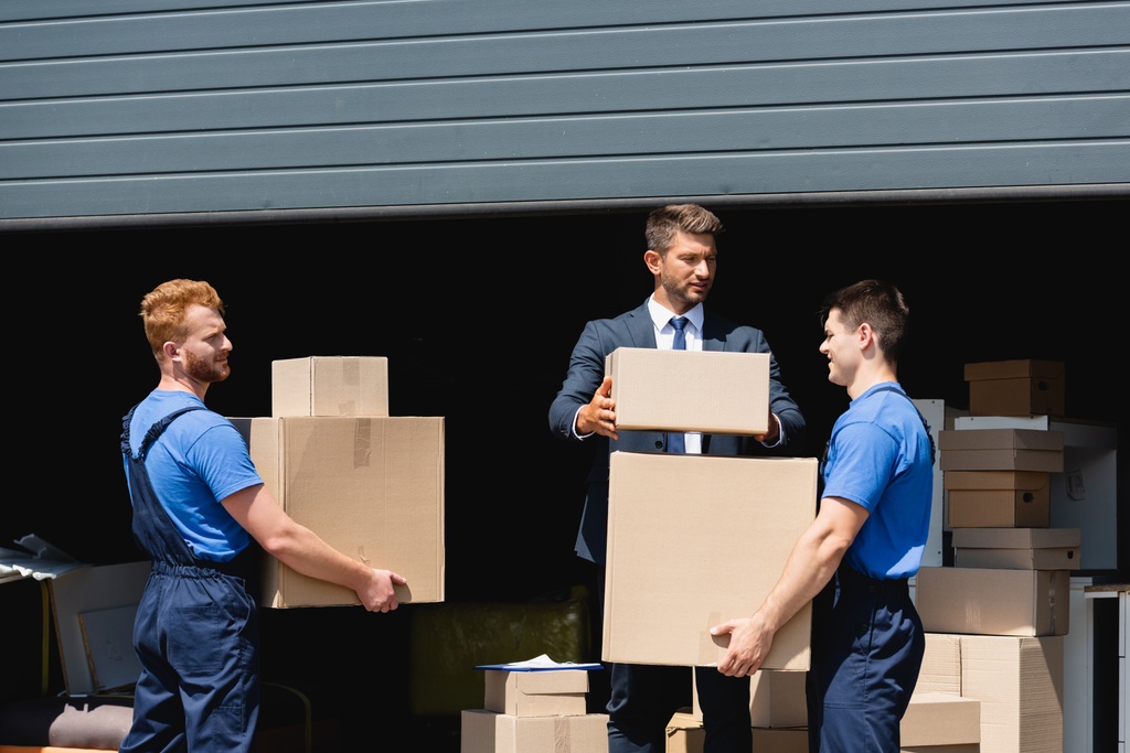 businessman in formal wear stacking packages near movers working outdoors - Photo, Image