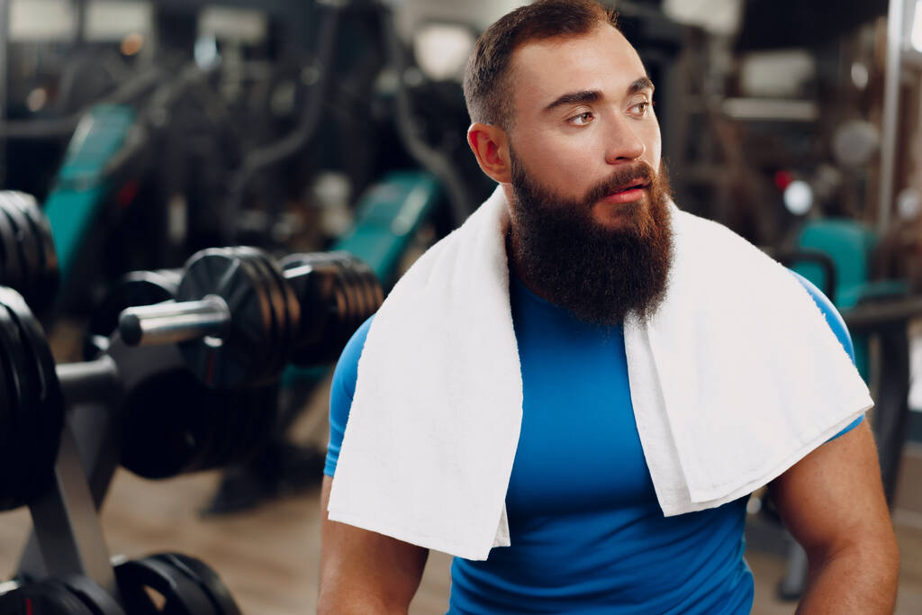 Бородатый молодой человек культурист в синей футболке, стоящий в спортзале - Фото, изображение