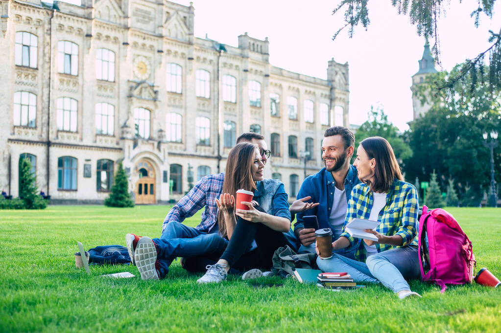 Щаслива красива команда студентів з рюкзаками та іншими речами розмовляє, розважається та відпочиває на траві на фоні коледжу на відкритому повітрі
 - Фото, зображення