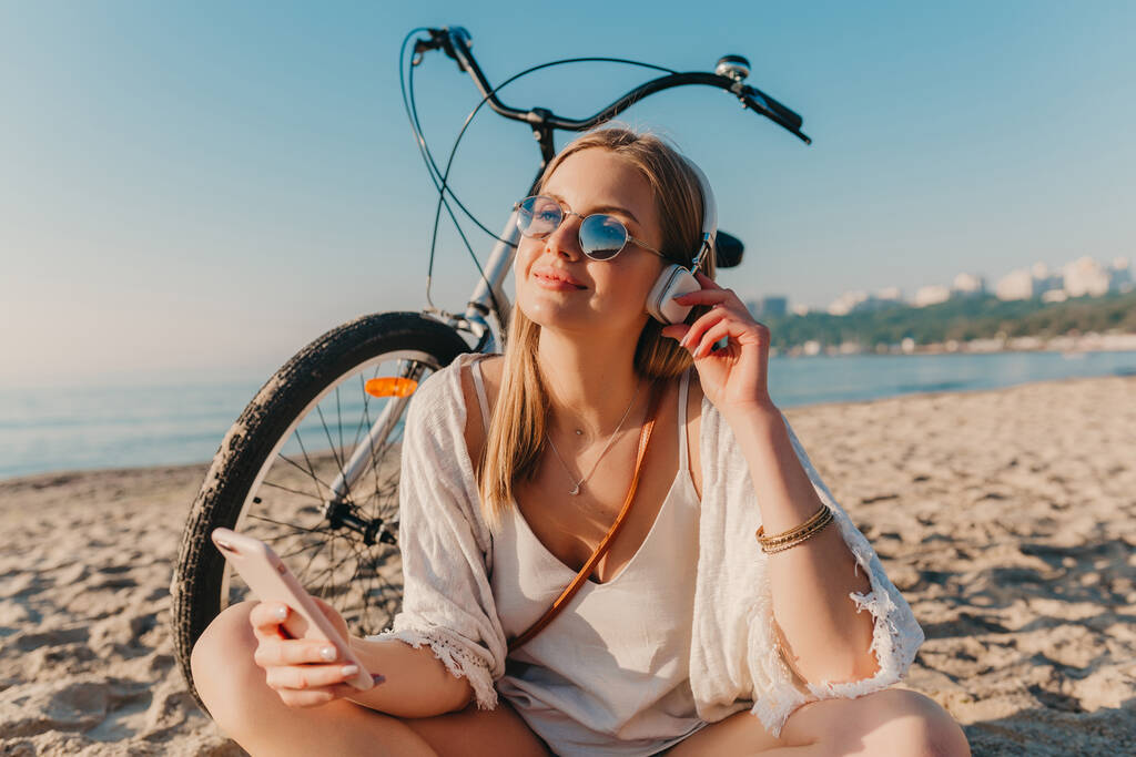 elegante joven atractiva rubia sonriente mujer sentada en la playa con bicicleta en auriculares escuchando música relajado usando el teléfono - Foto, Imagen