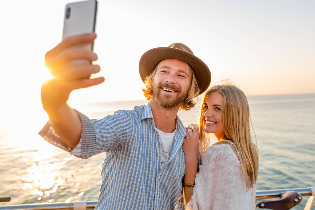 jeune homme et femme heureux souriant attrayant voyageant sur des vélos prenant selfie photo sur la caméra du téléphone, couple romantique au bord de la mer au coucher du soleil, tenue de style boho hipster, amis s'amuser ensemble - Photo, image