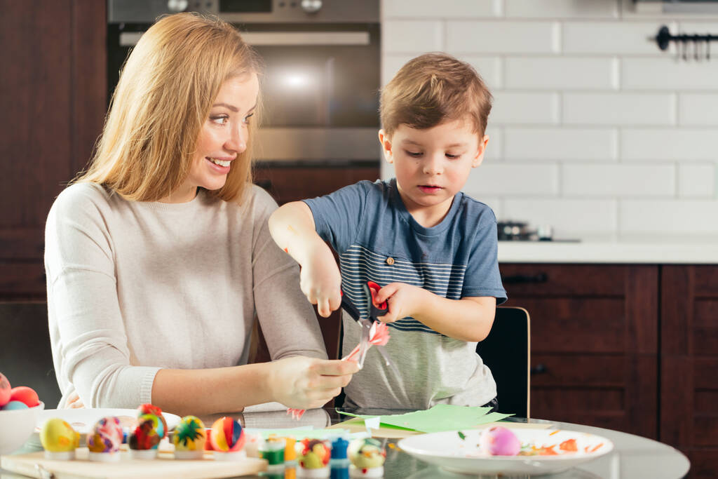 Mother and son cutting out paper with scissors at home, preparing Easter decor. - Photo, Image