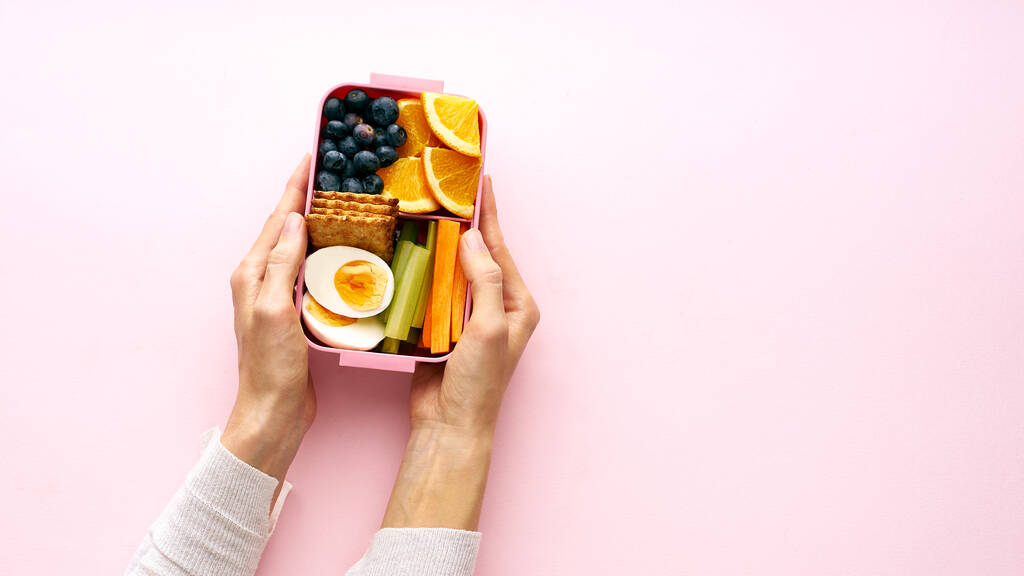 Жіночі руки тримають обідню коробку з яйцем та апельсиновими фруктами з чорницею та крекерами, вид зверху
 - Фото, зображення
