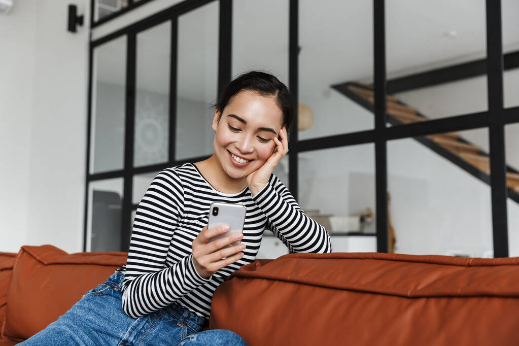 Ελκυστική χαμογελαστή νεαρή Ασιάτισσα που φοράει καθημερινά ρούχα χαλαρώνοντας σε έναν δερμάτινο καναπέ στο σπίτι, χρησιμοποιώντας κινητό τηλέφωνο - Φωτογραφία, εικόνα