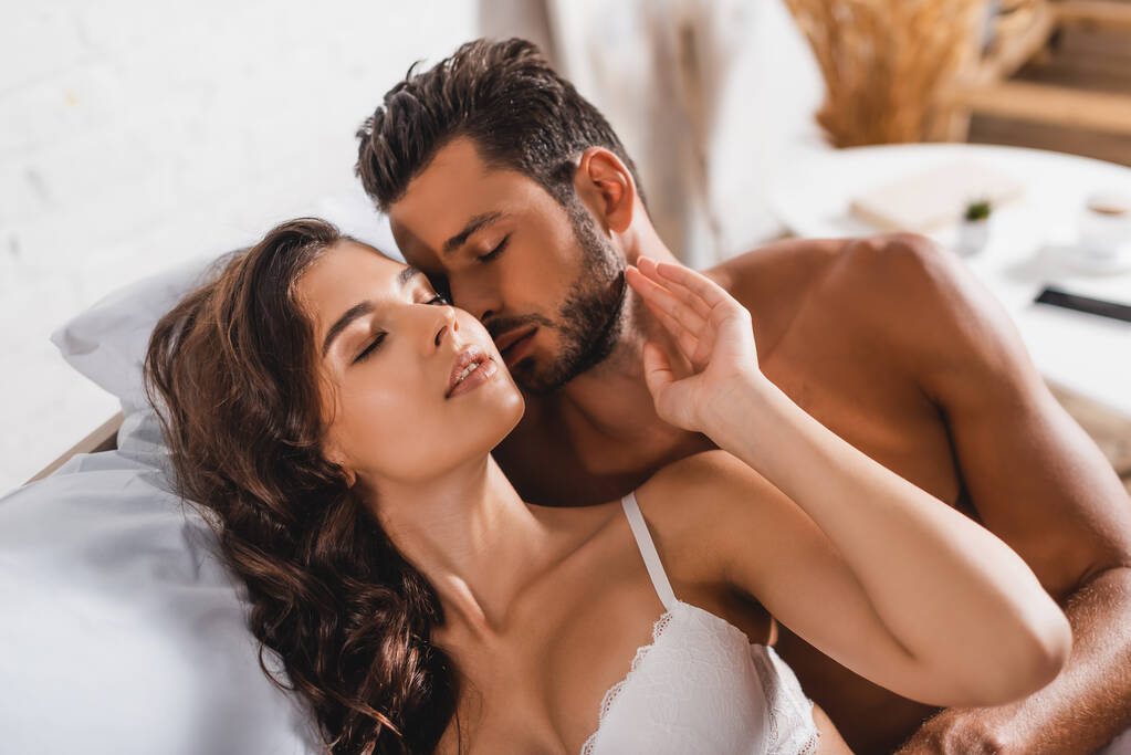 Hombre sin camisa besando a mujer sensual en sujetador con los ojos cerrados en la cama  - Foto, imagen