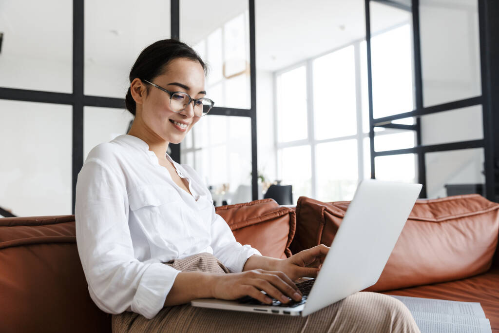 Attraente sorridente giovane donna d'affari asiatica che si rilassa su un divano di pelle a casa, lavorando sul computer portatile - Foto, immagini