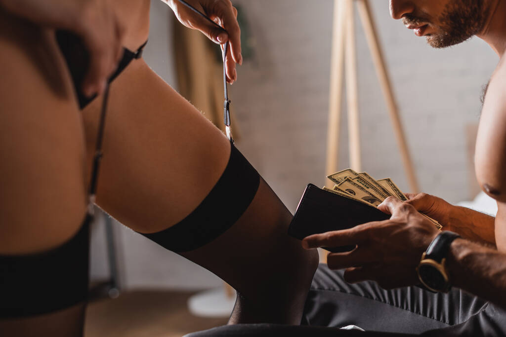 Valikoiva painopiste paidaton mies tilalla dollareita ja lompakko lähellä seksikäs nainen koskettaa sukkanauha vyö  - Valokuva, kuva