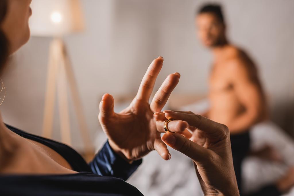Focus selettivo della donna che toglie la fede nuziale mentre si trova vicino all'uomo senza maglietta in camera da letto  - Foto, immagini