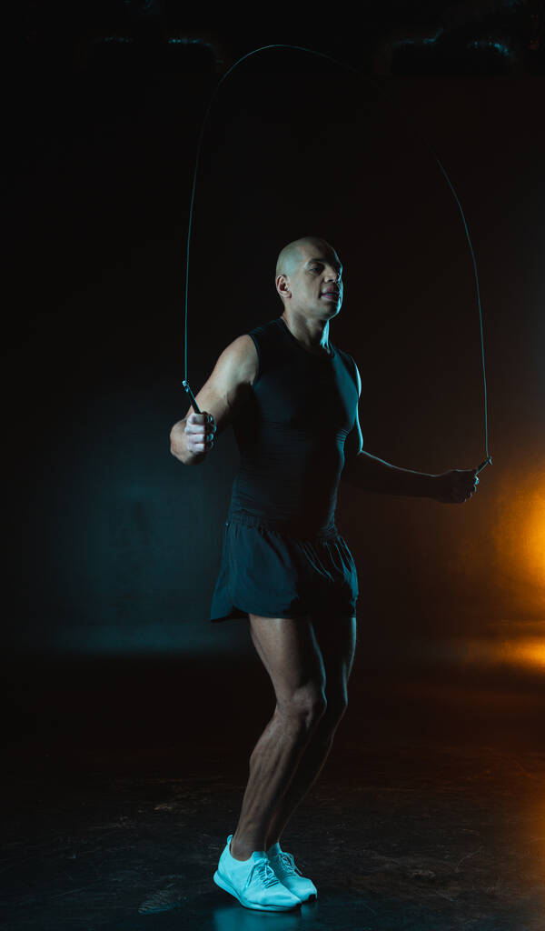 Esportista muscular confiante pulando em uma corda de salto dentro de casa contra o fundo escuro
 - Foto, Imagem
