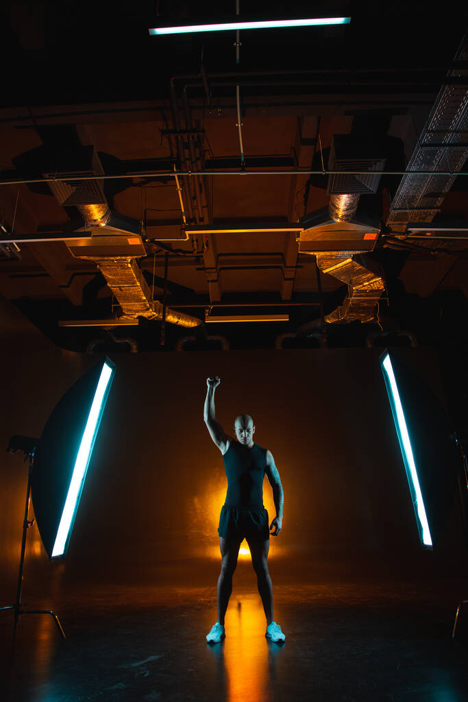 Esportista muscular confiante em pé no estúdio entre duas softboxes e colocando um punho para cima
 - Foto, Imagem