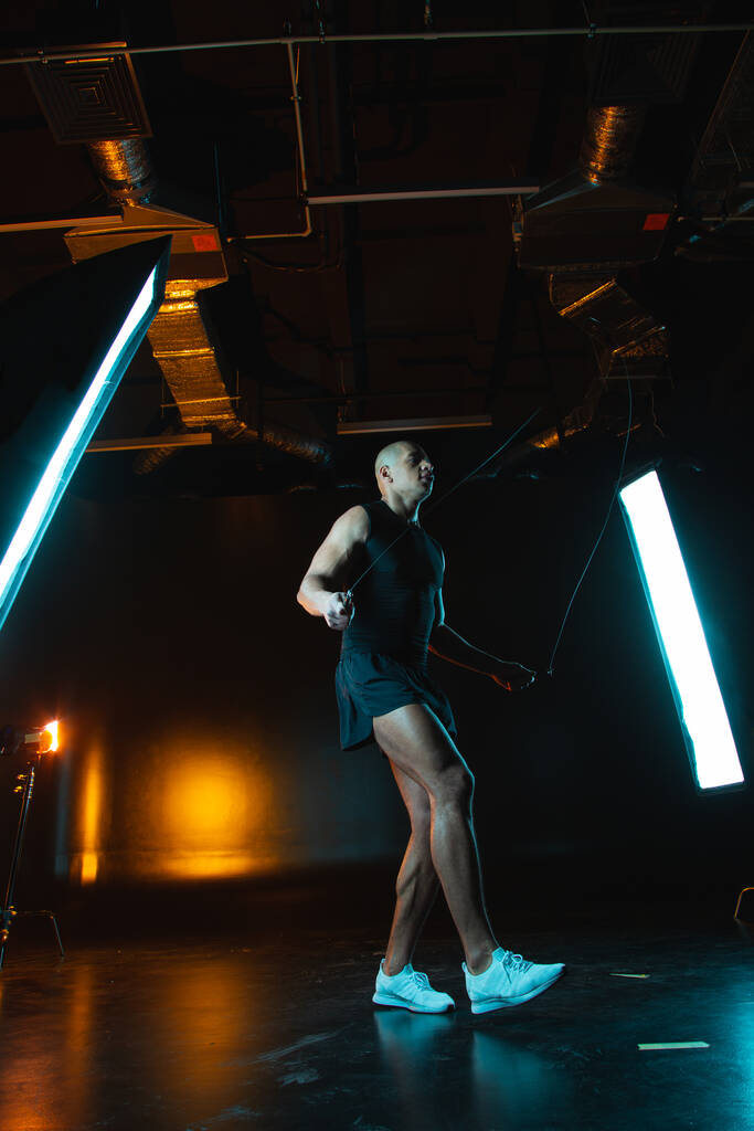 Atractivo hombre musculoso entrenando en una cuerda de saltar mientras está en el estudio oscuro con softboxes de luz a su alrededor - Foto, Imagen