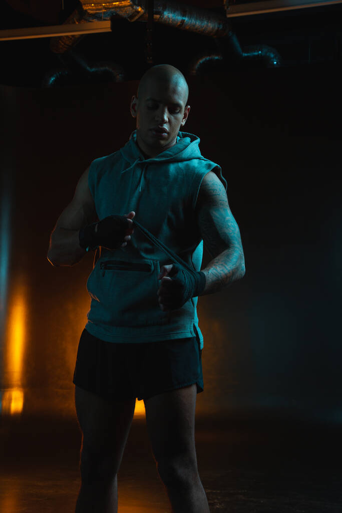 Klidný svalnatý muž hledá soustředěný, zatímco stojí v tmavém studiu a váže boxerské obvazy - Fotografie, Obrázek