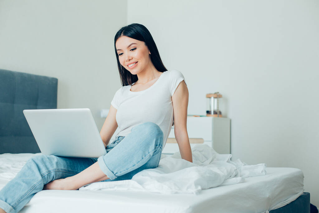 mooie vrouw glimlachen en kijken naar het scherm van een moderne laptop terwijl zitten op het bed met het - Foto, afbeelding