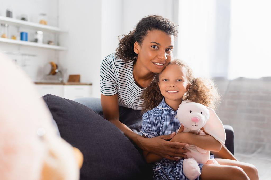 foco seletivo da mulher americana africana em camiseta listrada, e filha com coelho de brinquedo olhando para a câmera  - Foto, Imagem