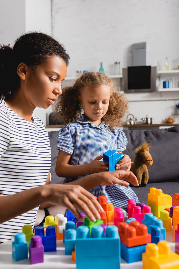 enfoque selectivo de niñera afroamericana en camiseta a rayas y niña en vestido jugando con bloques de construcción de colores en casa - Foto, imagen