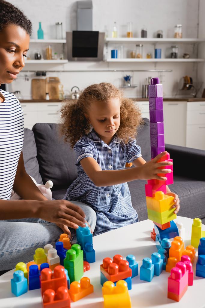 enfoque selectivo de la torre de construcción de niñas afroamericanas de bloques multicolores cerca de la niñera joven  - Foto, Imagen