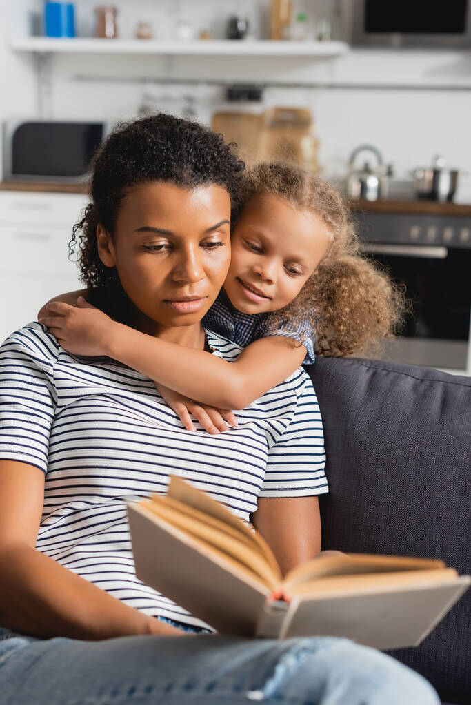 selectieve focus van Afrikaans Amerikaans meisje omarmen nanny in gestreept t-shirt lezen boek op bank - Foto, afbeelding