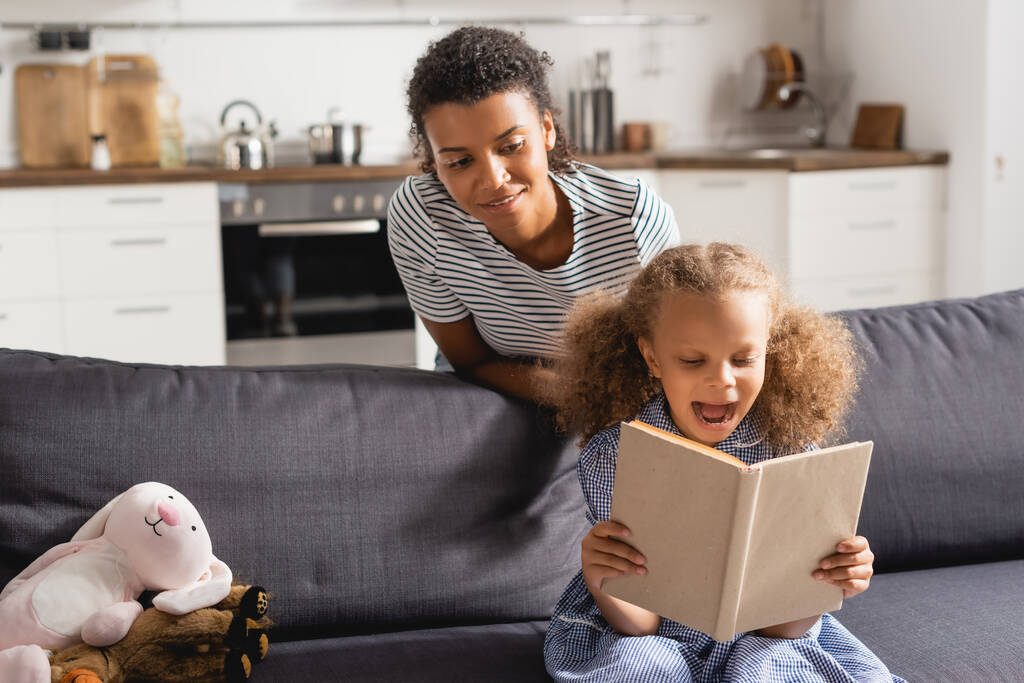 young african american babysitter behind excited child reding book on couch in kitchen - Photo, Image