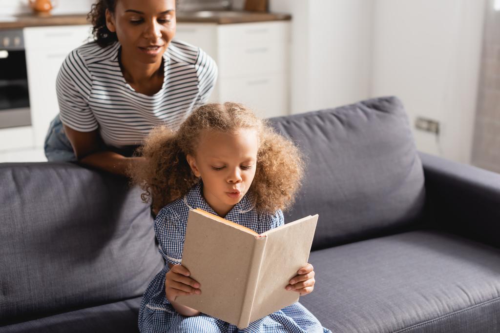 Αφροαμερικανή νταντά με ριγέ μπλουζάκι που κοιτάζει συμπυκνωμένο κορίτσι που κάθεται στον καναπέ και διαβάζει βιβλίο. - Φωτογραφία, εικόνα