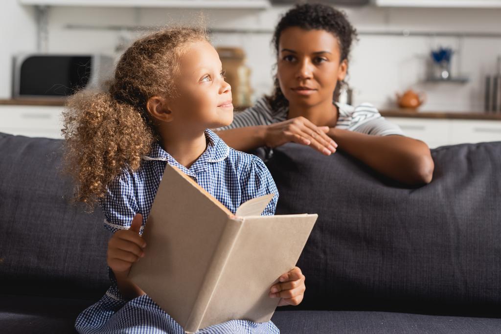 enfoque selectivo de niñera afroamericana detrás de niño soñador mirando hacia otro lado mientras sostiene libro abierto - Foto, Imagen