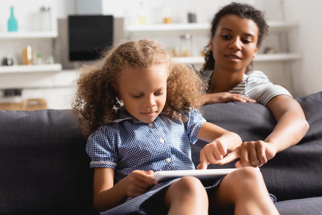 selective focus of african american nanny pointing with finger at digital tablet in hands of child sitting on sofa - Photo, Image