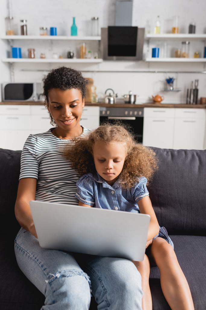 jovem freelancer afro-americano trabalhando no laptop enquanto se senta com a filha no sofá na cozinha - Foto, Imagem