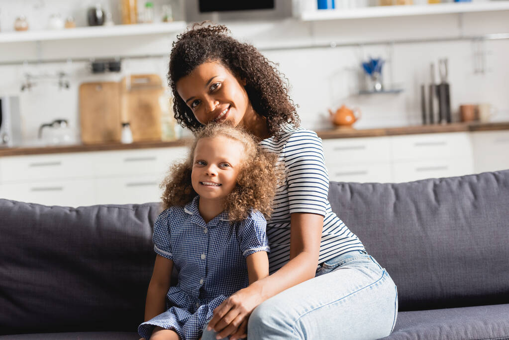 african american mother and daughter sitting on couch in kitchen and looking at camera - Photo, Image