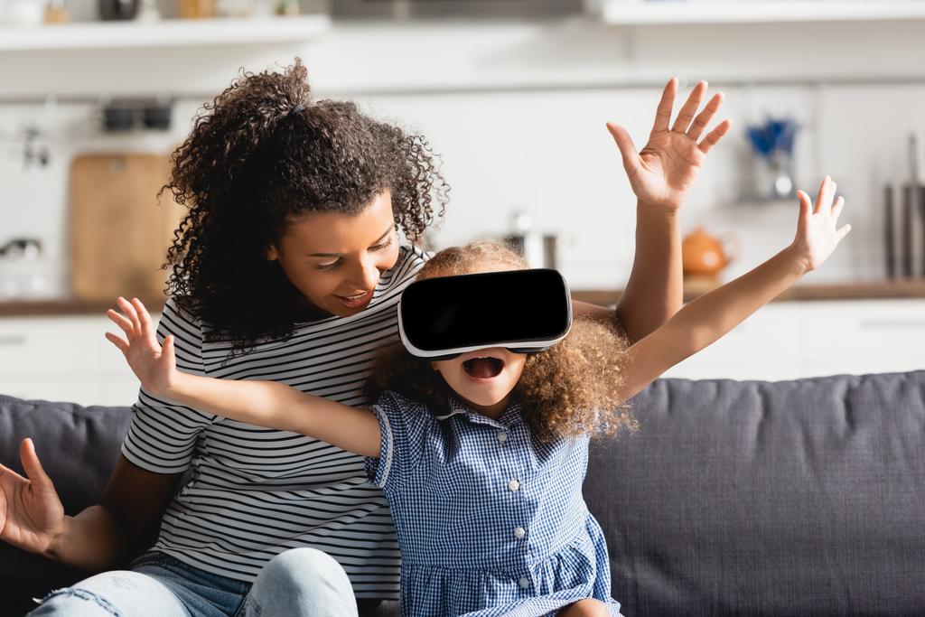 junge afrikanisch-amerikanische Kindermädchen und aufgeregtes Mädchen in vr Headset gestikulierend mit ausgestreckten Händen auf der Couch - Foto, Bild