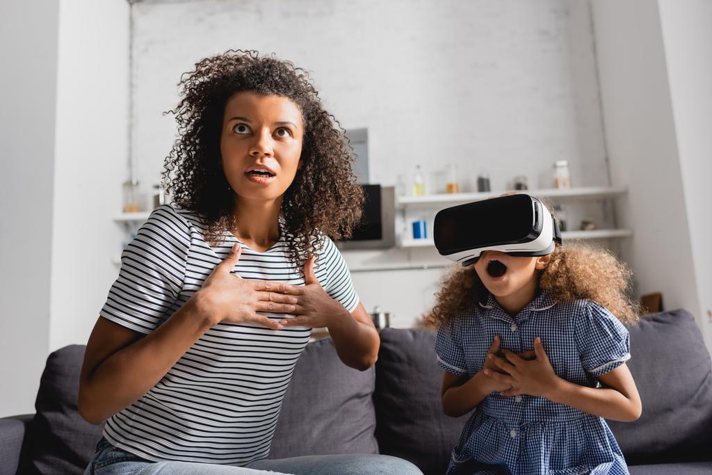 excited african american nanny and girl in vr headset holding hands on chests while sitting with open mouths - Photo, Image