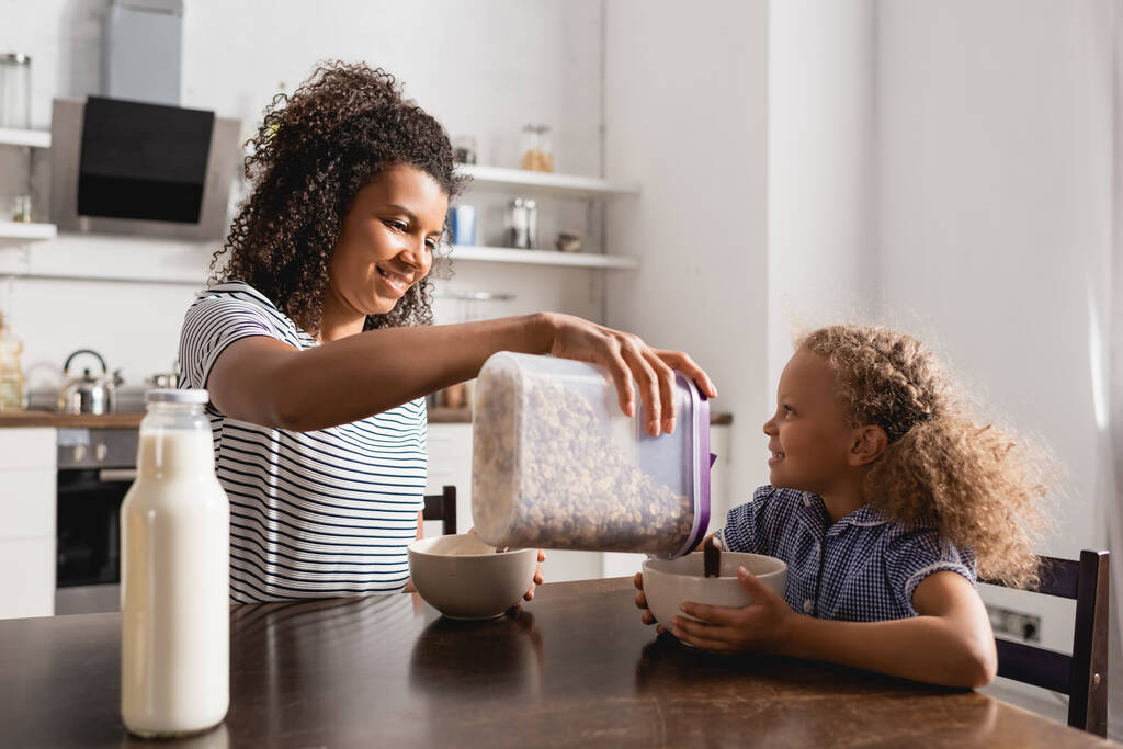 Afrikanisch-amerikanische Mutter in gestreiftem T-Shirt gießt Cornflakes in Schüssel neben Tochter und Flasche frischer Milch - Foto, Bild