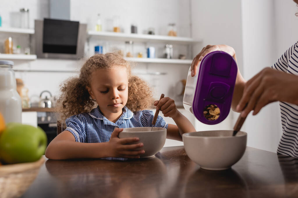 częściowy widok afrykańskiej amerykanki mama wylewanie płatków kukurydzianych do miski w pobliżu córki jedząc śniadanie - Zdjęcie, obraz