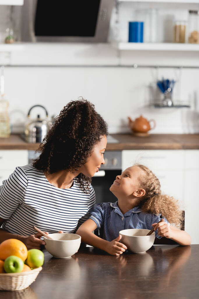 afro-américaine mère et fille regardant l'autre tout en étant assis près des bols avec le petit déjeuner - Photo, image