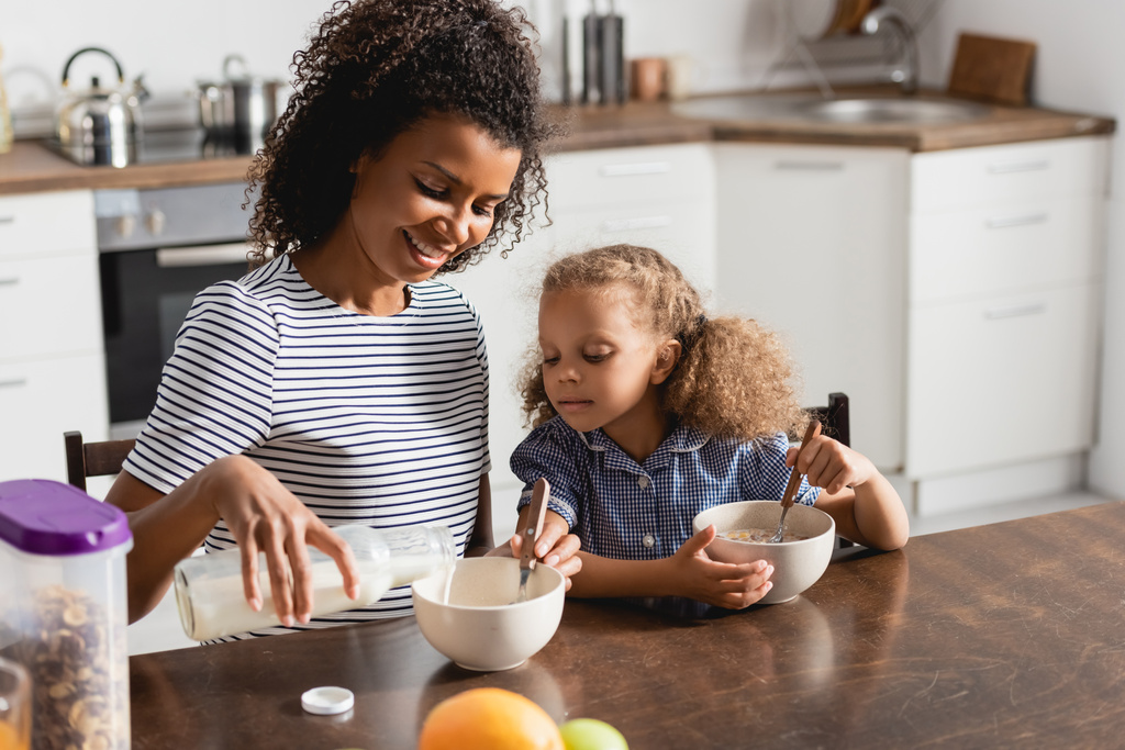 maman américaine africaine en t-shirt rayé versant du lait dans un bol tout en prenant le petit déjeuner avec sa fille - Photo, image