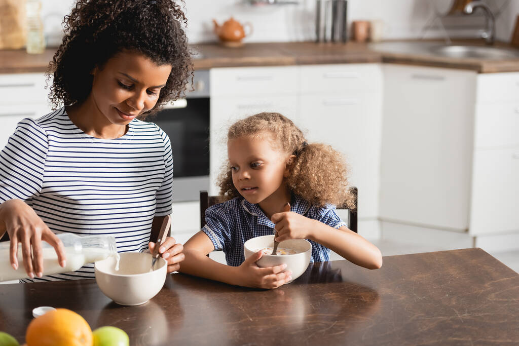 молодая африканская американка в полосатой футболке, наливающая молоко в миску во время завтрака с дочерью - Фото, изображение
