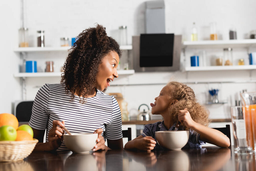 podekscytowany Afroamerykanka kobieta w paski t-shirt patrząc na córkę wystające język podczas śniadania w kuchni - Zdjęcie, obraz