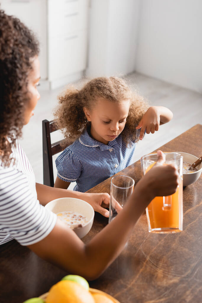 enfoque selectivo de la madre sosteniendo jarra con jugo de naranja cerca de hija afroamericana señalando con el dedo durante el desayuno - Foto, imagen