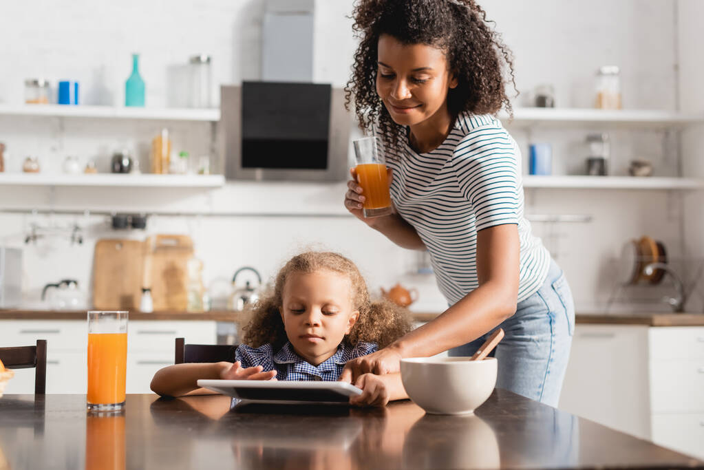 africká americká matka drží pomerančovou šťávu a dotýká digitální tablet v blízkosti koncentrovaného dítěte v kuchyni - Fotografie, Obrázek
