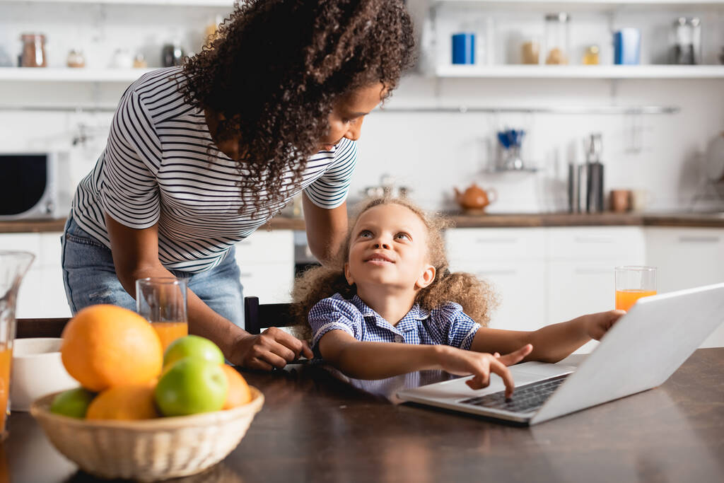 enfoque selectivo de la madre y la hija afroamericana mirándose cerca de la computadora portátil en la cocina - Foto, imagen