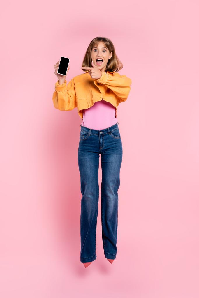 Ενθουσιασμένη γυναίκα άλμα και δείχνοντας με το δάχτυλο στο smartphone σε ροζ φόντο - Φωτογραφία, εικόνα