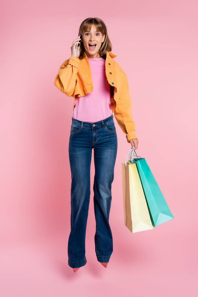 Mujer emocionada sosteniendo bolsas de compras y hablando en el teléfono inteligente mientras salta sobre fondo rosa - Foto, imagen
