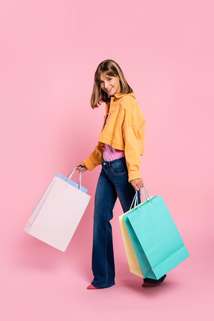 Nő sárga dzsekiben nézi a kamerát, miközben kezében bevásárló táskák rózsaszín háttér - Fotó, kép