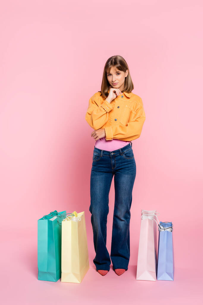 Jovem mulher olhando para a câmera perto de sacos de compras coloridos no fundo rosa - Foto, Imagem