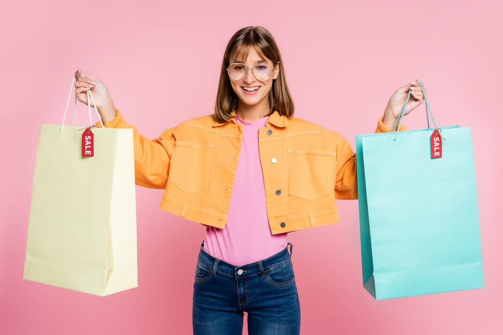 Young woman in sunglasses looking at camera while holding colorful shopping bags with sale word on price tags on pink background - Photo, Image