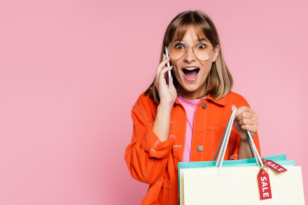 Shocked woman looking at camera while holding purchases with sale word on price tags and talking on smartphone on pink background - Photo, Image